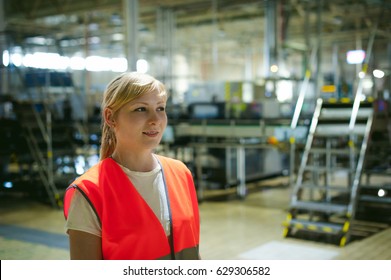 Portrait Of A Female Employee In An Orange Robe Vest In The Working Space Of A Production Facility, Supervises And Controls The Line In Food Production, Against The Background Of Equipment