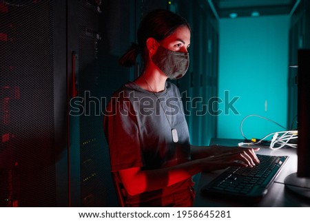 Portrait of female IT engineer wearing mask while using computer and working in server room lit by red light, copy space
