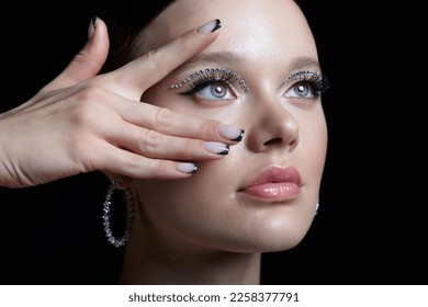 Portrait of female face with unusual rhinestones makeup. Woman with earring in the form of a shiny ring in the ear. Girl with nails with black shiny manicure. Hand near face.