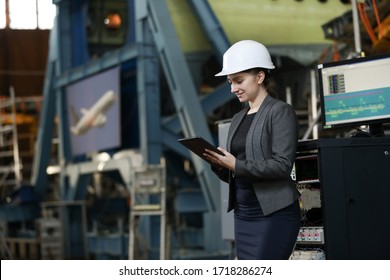 Portrait Of A Female Factory Manager In A White Hard Hat And Business Suit. Controlling The Work Process At The Airplane Manufacturer.