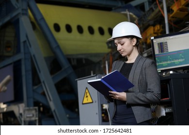 Portrait Of A Female Factory Manager In A White Hard Hat And Business Suit. Controlling The Work Process At The Airplane Manufacturer.