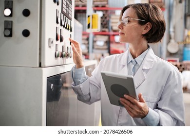 Portrait Of Female Factory Worker Wearing Glasses And White Robe, Pressing Machine Buttons, Using Tablet For Quality Control And Logistic Purposes At Polymer Plastic Manufacturing