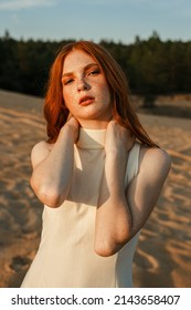Portrait of female fashion model with red hair and freckles touching neck on sand in nature in summer evening while looking at camera 