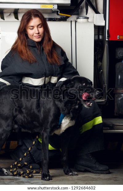 Portrait of female firefighter with black dog\
sitting on background of fire\
truck
