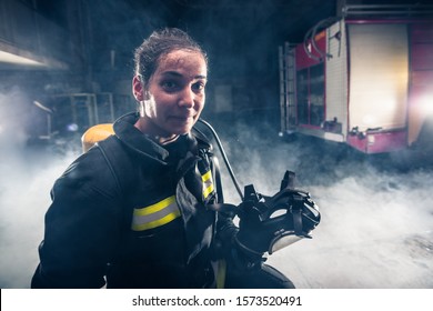 Portrait Of A Female Firefighter While Holding An Axe And Wearing An Oxygen Mask Indoors Surrounded By Smoke.