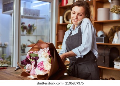 Portrait Of Female Florist In Apron Arranging Fresh Flowers For Bouquet In The Flower Shop, Using Roses, Hydrangea, Peonies, And Holding Cell Phone With Shoulder, Busy On The Call