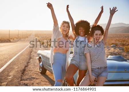 Portrait Of Female Friends Enjoying Road Trip Standing Next To Classic Car On Desert Highway