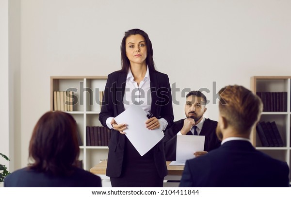 Portrait of female lawyer or attorney who\
represents plaintiff in lawsuit during court trial. Serious\
unsmiling woman holding documents standing in courtroom after her\
speech in front of\
audience