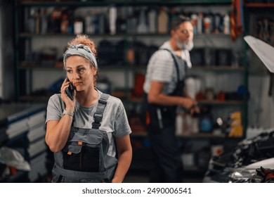 Portrait of female mechanic standing at car repair shop and having phone conversation with customer and looking away. In a blurry background is male mechanic fixing car under the hood at workshop. - Powered by Shutterstock