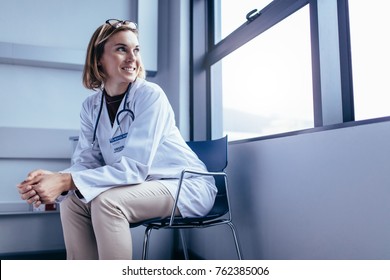 Portrait Of Female Medical Professional Sitting On Chair In Hospital Room. Doctor In Hospital Ward Looking Outside The Window.