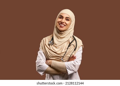 Portrait of a female Muslim doctor wearing a white coat, arms crossed, standing against a brown background and smiling. The concept of medicine, care, health. - Shutterstock ID 2128112144