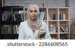 Portrait of female muslim student, wearing hijab, who is at campus library and confidently looking at camera - modern Islam concept 