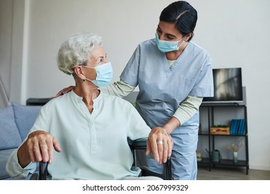 Portrait Of Female Nurse Assisting Senior Woman In Wheelchair At Home Or In Retirement Center, Both Wearing Masks