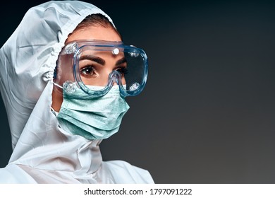 Portrait Of Female Nurse In Medical Mask With Safety Glasses And In White Protective Suit On Grey Background. Side View. Copy Space