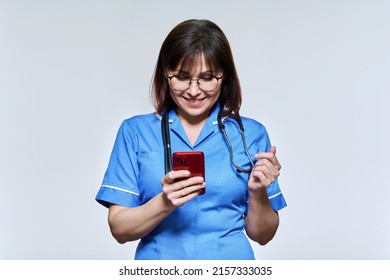 Portrait Of Female Nurse With Smartphone In Hands, Looking At Phone On Light Background