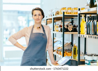 Portrait of female owner saleswoman in grey apron selling products in zero waste shop. Female owner weighing oil in glass bottle in plastic free grocery store. Eco shopping at local business concept. - Powered by Shutterstock