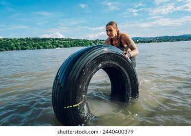 Portrait of a female participating in an obstacle course race outdoor. Wearing moisture-wicking clothing and gloves for protection while pushing tractor tire in a shallow water. Copy space. - Powered by Shutterstock