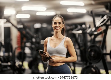 Portrait of a female personal trainer standing in a gym with tablet in her hands and smiling at the camera. - Powered by Shutterstock