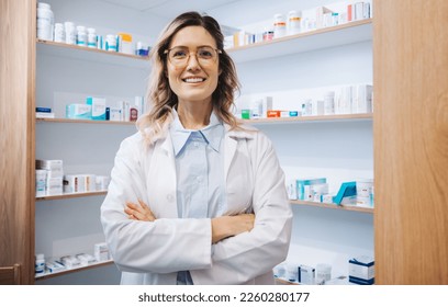 Portrait of a female pharmacist standing in a drug store. Female healthcare worker smiling at the camera with crossed arms. Woman working in a pharmacy. - Powered by Shutterstock