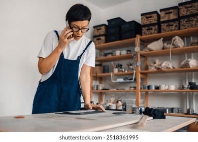 Portrait of female pottery designer in overalls standing at ceramic studio and having a phone conversation. Young artistic creative female ceramist working with wet clay and talking on her cellphone. - Powered by Shutterstock