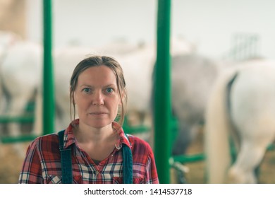 Portrait Of Female Rancher At Horse Stable Looking At Camera. Adult Woman Wearing Plaid Shirt And Jeans Bib Overalls As Farm Worker.