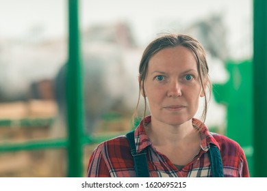 Portrait Of Female Rancher At Horse Stable Looking At Camera. Adult Woman Wearing Plaid Shirt And Jeans Bib Overalls As Farm Worker.