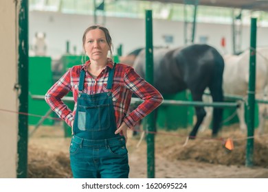 Portrait Of Female Rancher At Horse Stable Looking At Camera. Adult Woman Wearing Plaid Shirt And Jeans Bib Overalls As Farm Worker.