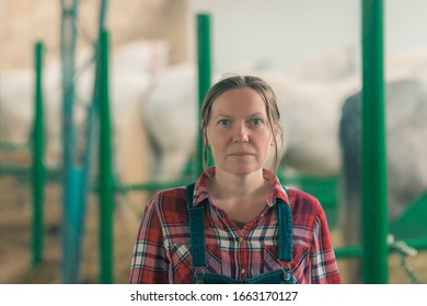 Portrait Of Female Rancher At Horse Stable Looking At Camera. Adult Woman Wearing Plaid Shirt And Jeans Bib Overalls As Farm Worker.