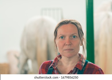 Portrait Of Female Rancher At Horse Stable Looking At Camera. Adult Woman Wearing Plaid Shirt And Jeans Bib Overalls As Farm Worker.