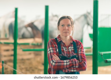 Portrait Of Female Rancher At Horse Stable Looking At Camera. Adult Woman Wearing Plaid Shirt And Jeans Bib Overalls As Farm Worker.