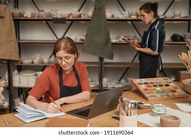 Portrait of female small business owner taking notes in planner while working at wooden table with laptop in pottery workshop, copy space - Powered by Shutterstock