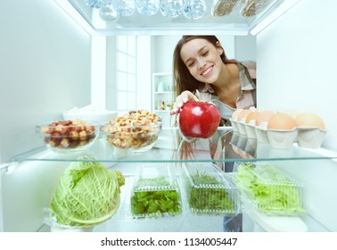 Portrait of female standing near open fridge full of healthy food, vegetables and fruits.