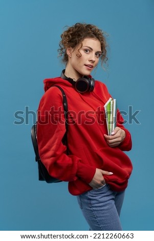 Portrait of female student posing, standing with books in hands. Young beautiful woman with rucksack studying, smiling, looking at camera. Isolated on blue studio background.