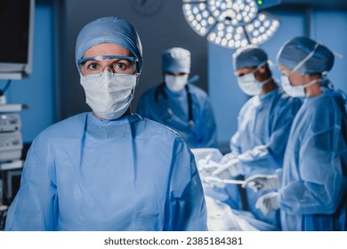 Portrait of female surgeon wearing scrubs and protective glasses in hospital operating theater. Serious surgeon doctor looking at camera in protective blue gown. Medical workers teamwork - Powered by Shutterstock