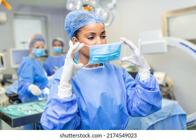 Portrait Of Female Surgeon Wearing Surgical Mask And Cap. Skilled Doctor Is Working In Illuminated Operating Room. She Is In Hospital.