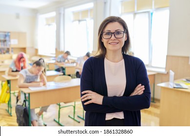Portrait of female teacher at school in class, smiling middle aged woman wearing glasses with folded hands looking at camera, classroom with teenage students at desk background - Powered by Shutterstock