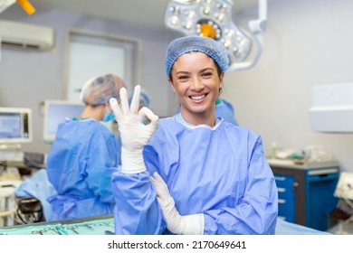 Portrait of female woman nurse surgeon OR staff member dressed in surgical scrubs gown mask and hair net in hospital operating room theater making eye contact smiling showing ok sign