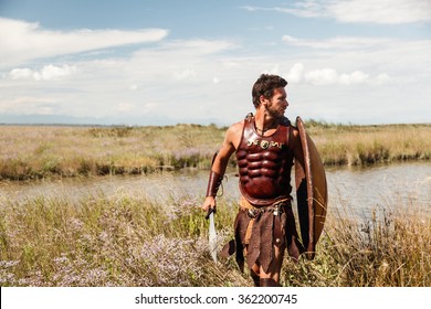 Portrait Of Fighting Ancient Warrior In Armor With Sword And Shield.  Spartan Soldier. Landscape Background