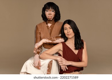 Portrait Of A Filipino Woman In Her 50's And An African American Woman With Vitiligo In Her 30's Sitting Confidently On A Neutral Background.