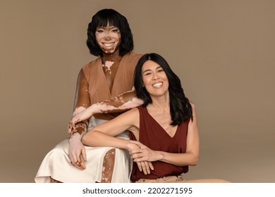 Portrait Of A Filipino Woman In Her 50's And An African American Woman With Vitiligo In Her 30's Sitting Together And Smiling On A Neutral Background.