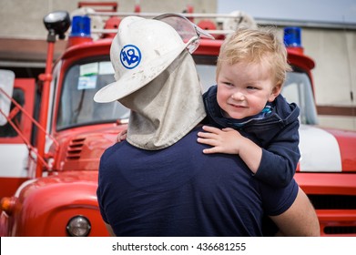 Portrait Of A Fireman Holding A Little Child Boy To Save Him. Fireman With Kid In Arms. Protection Concept. Fire Fighters And Rescuers Department Trucks On Fire Station. Protection Of Life