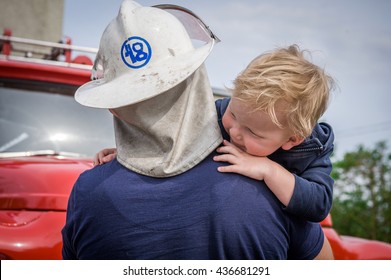 Portrait Of A Fireman Holding A Little Child Boy To Save Him. Fireman With Kid In Arms. Protection Concept. Fire Fighters And Rescuers Department Trucks On Fire Station. Protection Of Life