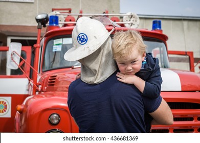 Portrait Of A Fireman Holding A Little Child Boy To Save Him. Fireman With Kid In Arms. Protection Concept. Fire Fighters And Rescuers Department Trucks On Fire Station. Protection Of Life