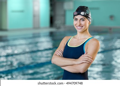 Portrait of a fit swimmer woman at pool looking at camera. Portrait of competitive female swimmer near swimming pool. Young swimmer wearing swim cap and goggles with crossed arms at poolside.