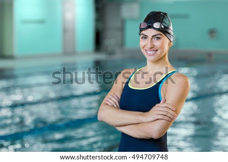 Portrait of a fit swimmer woman at pool looking at camera. Portrait of competitive female swimmer near swimming pool. Young swimmer wearing swim cap and goggles with crossed arms at poolside.
