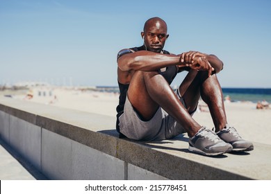 Portrait of fit young athlete relaxing on sea wall looking at camera. Muscular african man resting after his workout.
