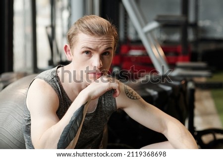 Portrait of fit young sportsman sitting on bench in gym, raising eyebrowns and looking at camera