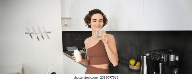 Portrait of fitness girl, healthy woman standing in kitchen with pills, takes vitamins with glass of water for healthy wellbeing. Sportswoman holds food supplements. - Powered by Shutterstock