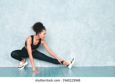 Portrait of fitness smiling black woman in sports clothing with afro curls hairstyle.She wearing sportswear.Young beautiful model stretching out before training.Female sitting in studio near gray wall