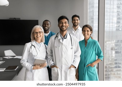 Portrait of five young and mature multiethnic medical professionals, therapists or GP wear uniforms standing together in clinic conference room, posing for camera. Hospital staff employees, mission - Powered by Shutterstock